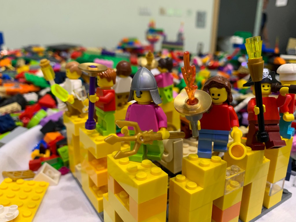 Lots of lego covering a table. In the foreground is a yellow castle with Lego figures defending the walls.